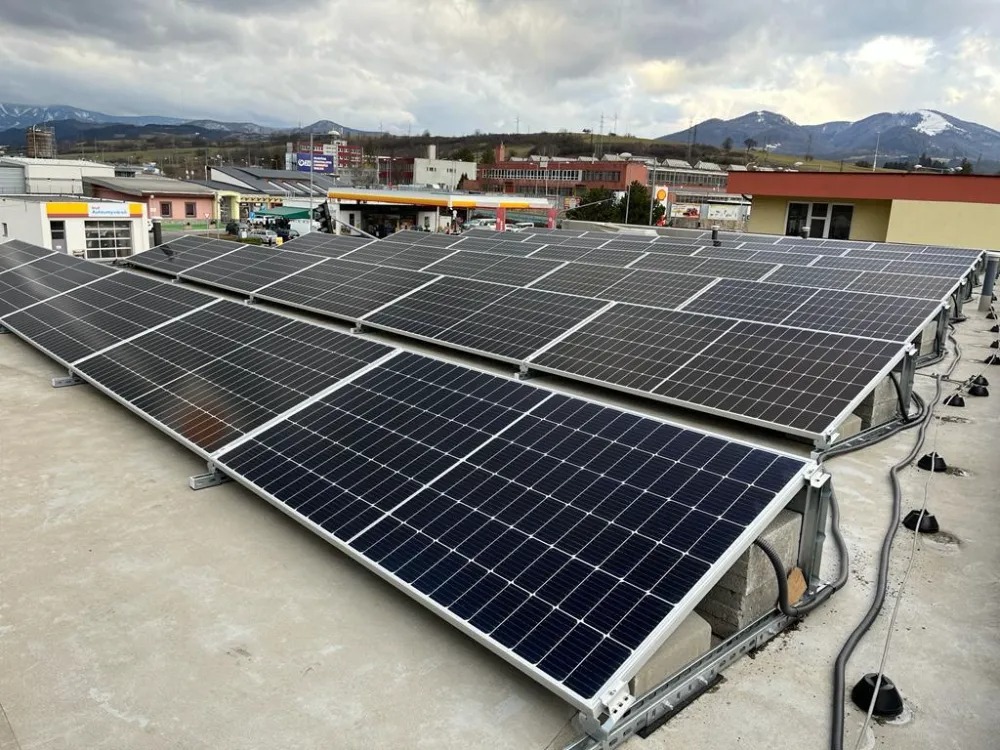 Rząd paneli słonecznych na płaskim dachu budynku z widokiem na zabudowania miejskie i góry w tle pod pochmurnym niebem.