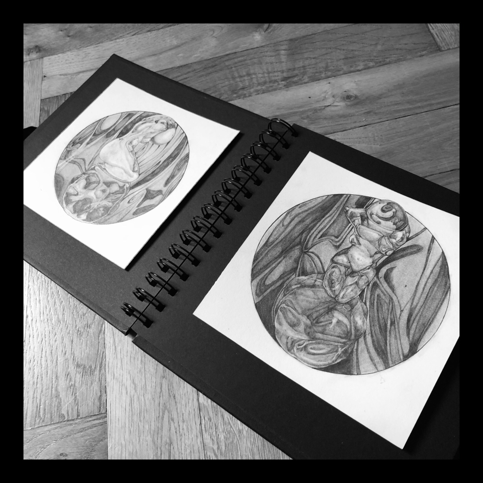 Szkicownik z dwoma rysunkami ołówkiem przedstawiającymi abstrakcyjne postacie w okręgach, leżący na drewnianej powierzchni.