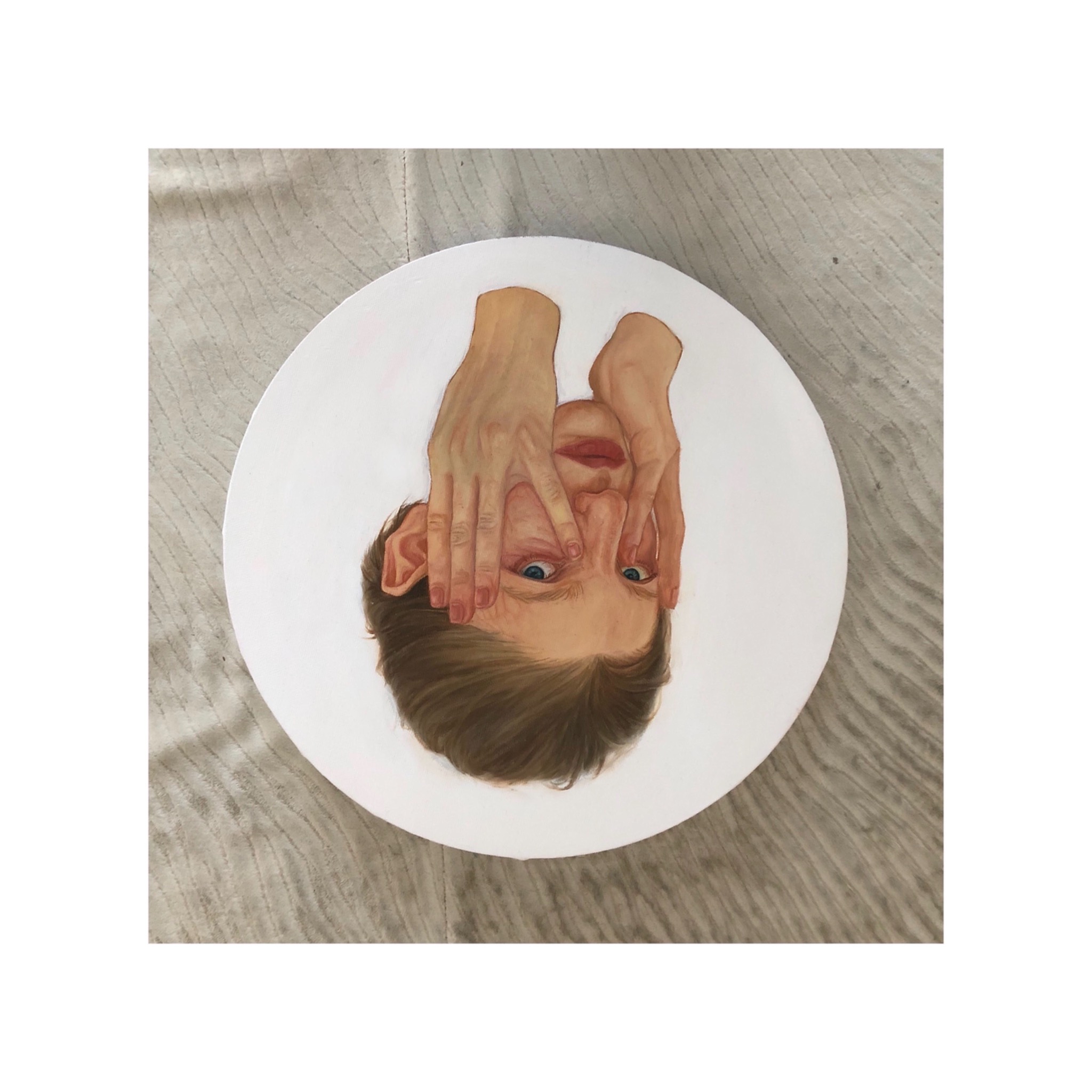 Okrągły obraz przedstawiający portret mężczyzny z niebieskimi oczami, którego twarz częściowo zasłaniają dłonie, leżący na teksturowanym materiale.