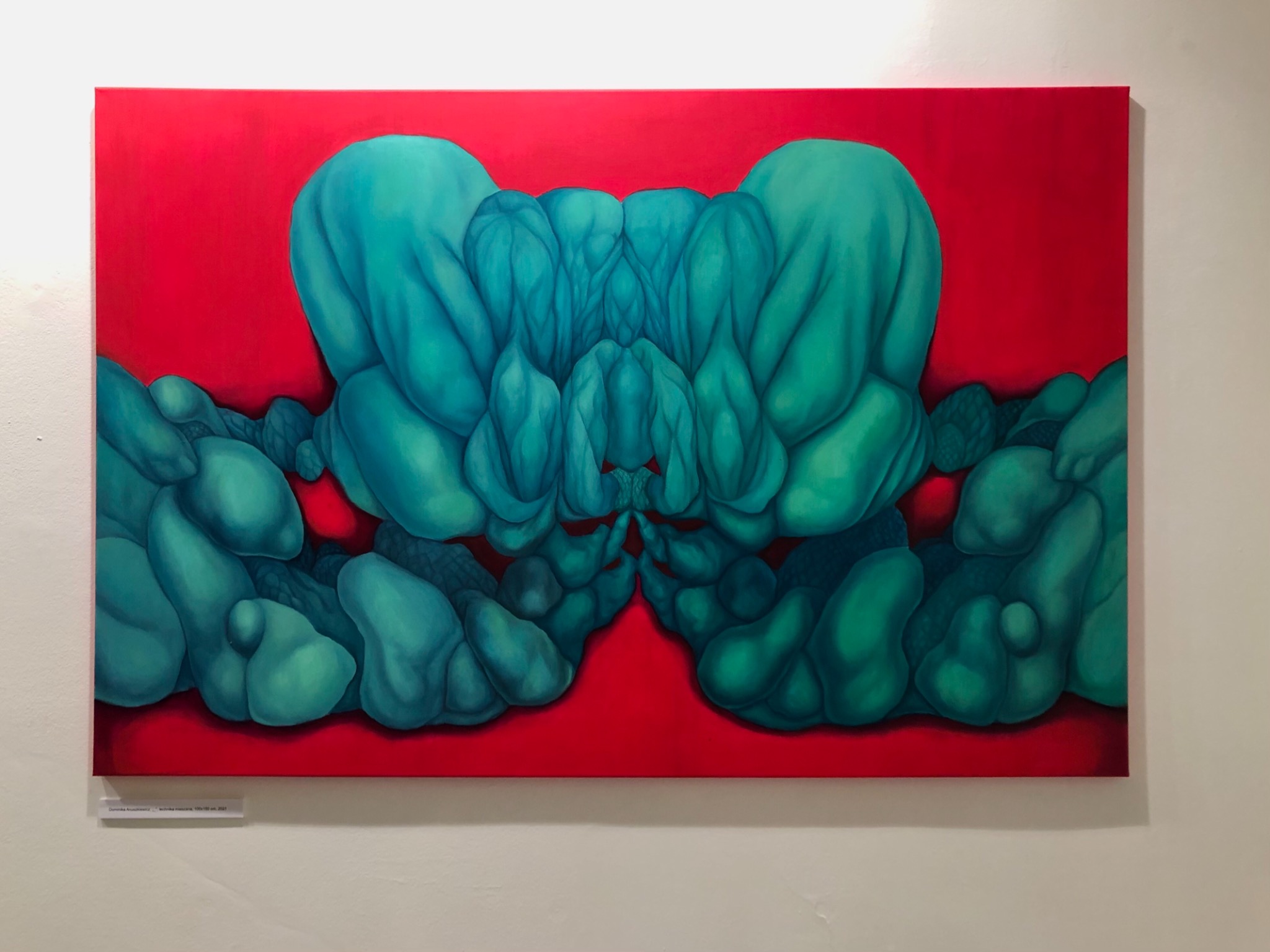 Abstrakcyjna kompozycja malarska z symetrycznym układem form w odcieniach turkusu i zieleni na intensywnie czerwonym tle, obraz zawieszony na białej ścianie.