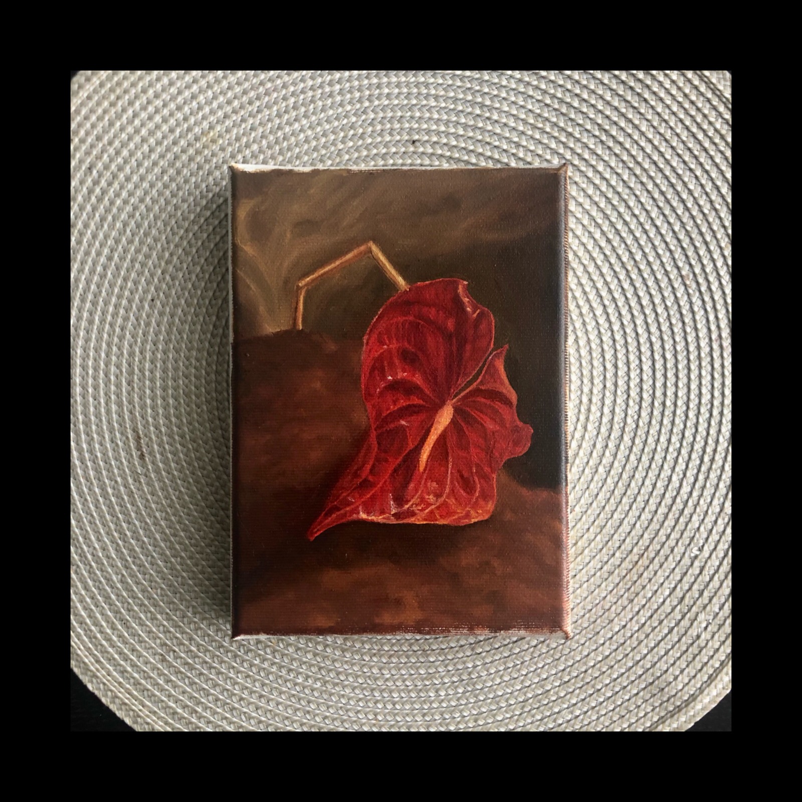 Olejny obraz na płótnie przedstawiający pojedynczy, czerwony kwiat anturium na ciemnobrązowym tle, z widoczną delikatną fakturą pędzla i metaliczną ramą w tle, umieszczony na szarej, okrągłej...
