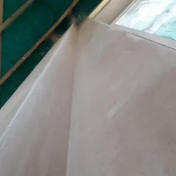 Ściana wewnętrzna po nałożeniu pierwszej warstwy szpachli, widoczna rama okienna i drewniana konstrukcja dachu z zieloną membraną, lekko nieostre ujęcie z dołu.