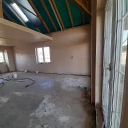 Surowe wnętrze z tynkami maszynowymi, widoczne narzędzia i materiały budowlane, okno dachowe w drewnianej konstrukcji dachu z zieloną membraną.