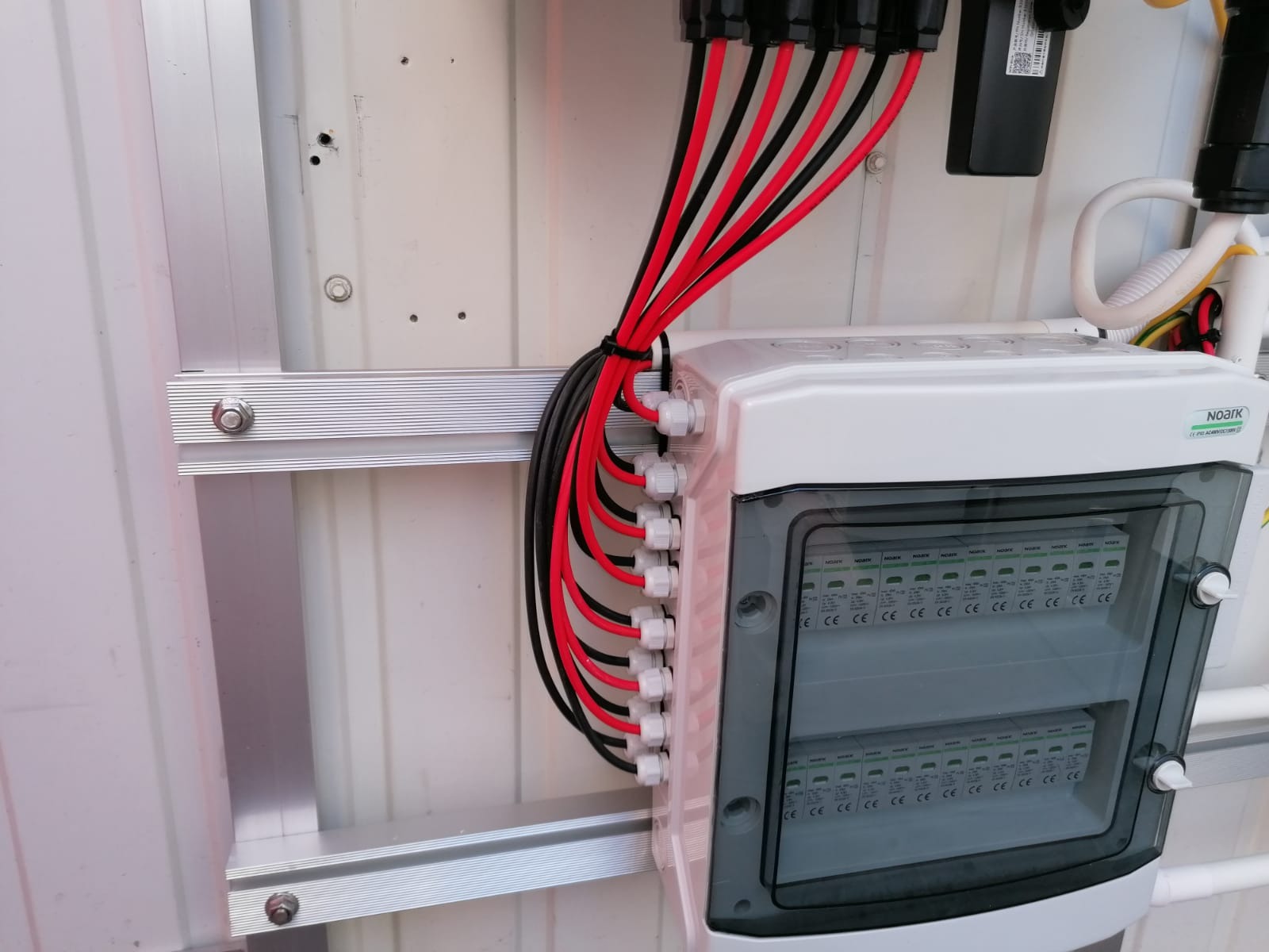 Rozdzielnica elektryczna z przejrzystą obudową, zamontowana na aluminiowej szynie, z podłączonymi kablami w kolorach czarnym i czerwonym, zabezpieczonymi białymi dławikami.