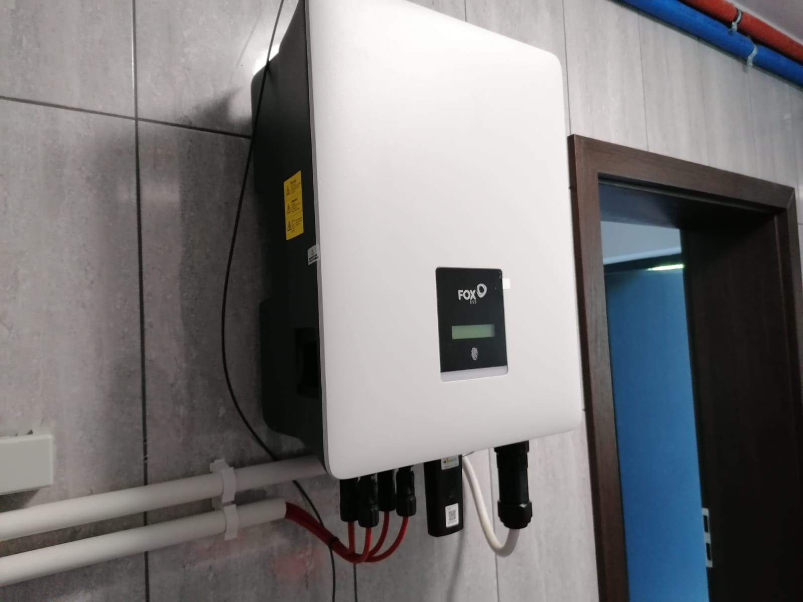 Biały inwerter fotowoltaiczny Fox ESS zamontowany na szarej, kafelkowanej ścianie wewnątrz budynku, z widocznymi przewodami i rurami instalacyjnymi.