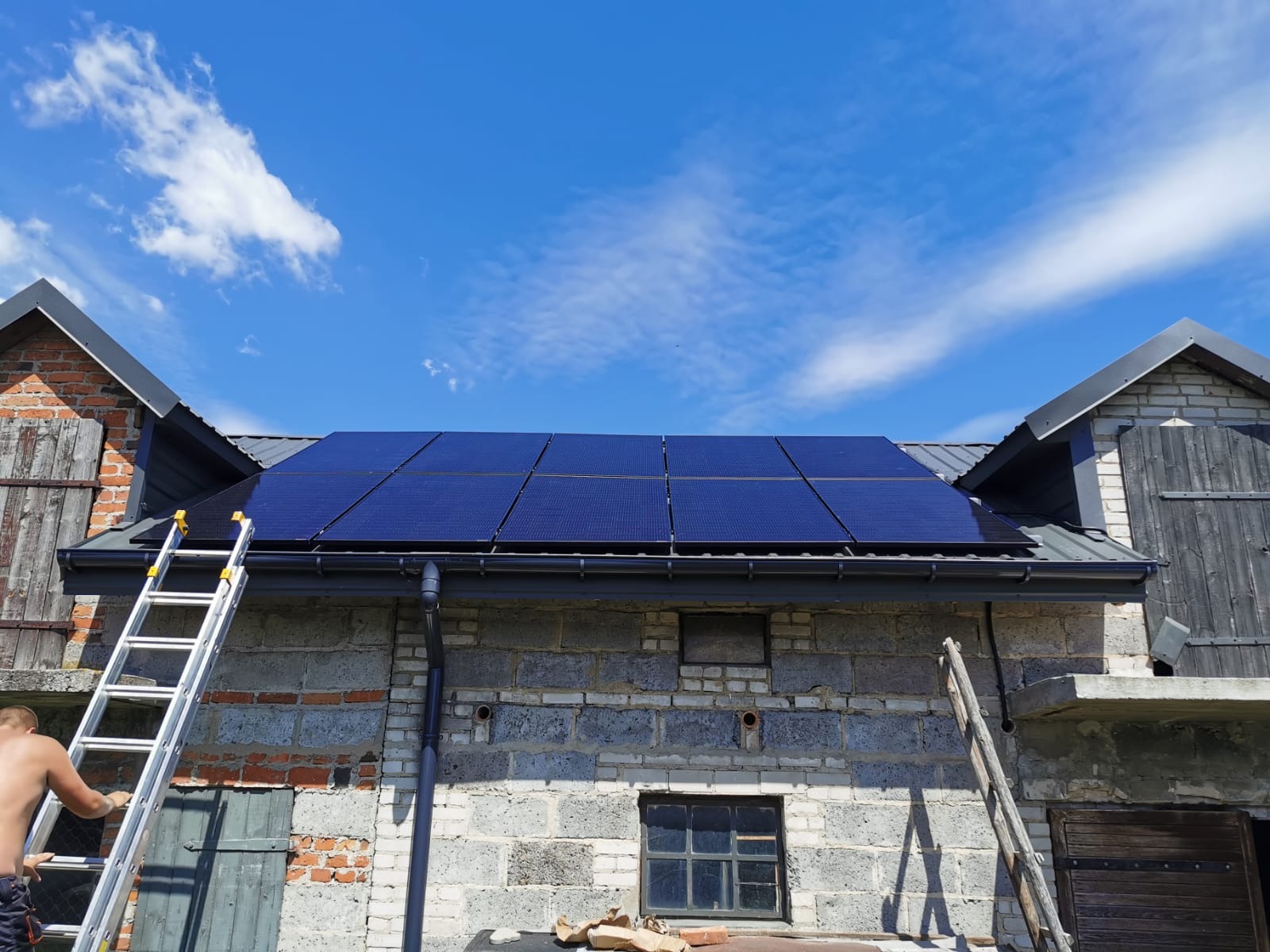 Montaż paneli na dachu budynku gospodarczego z cegły i pustaków, drabina oparta o ścianę, błękitne niebo z chmurami w tle.