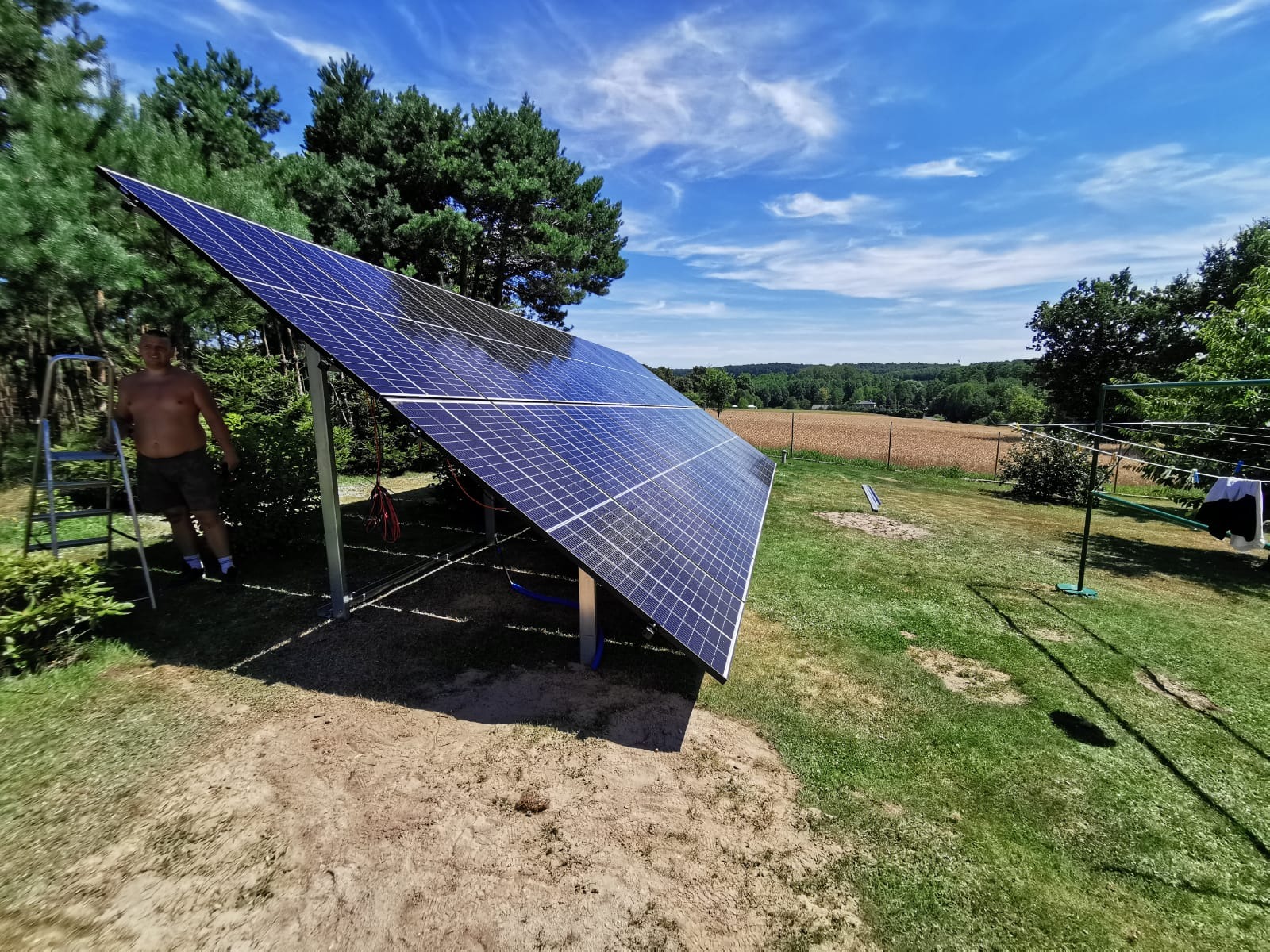 Montaż paneli słonecznych w ogrodzie, widoczny mężczyzna bez koszuli na drabinie, w tle pole i las pod błękitnym niebem.