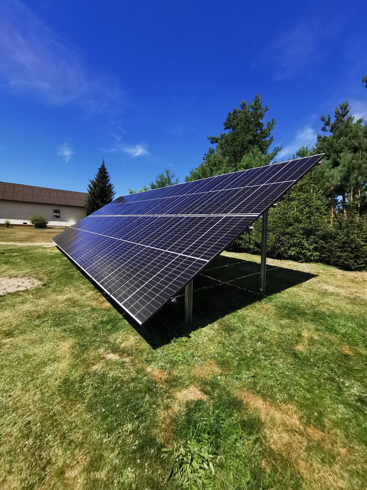 Panel słoneczny na metalowej konstrukcji ustawiony na trawniku przed budynkiem i drzewami w słoneczny dzień.