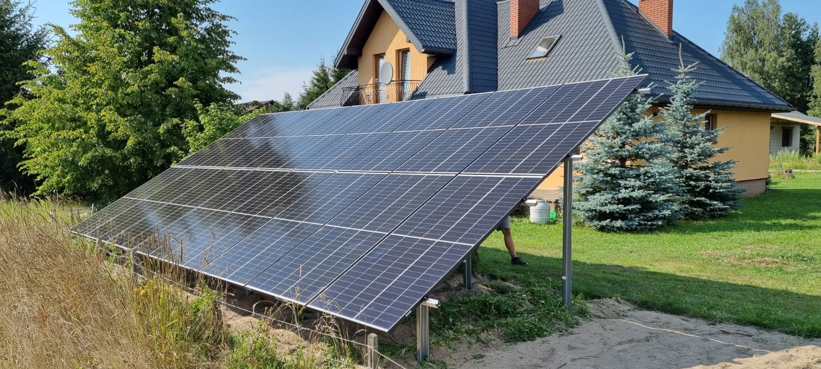 Instalacja paneli słonecznych na gruncie przed domem jednorodzinnym w słoneczny dzień. Widoczne panele, konstrukcja wsporcza, trawa, drzewa i fragment budynku.