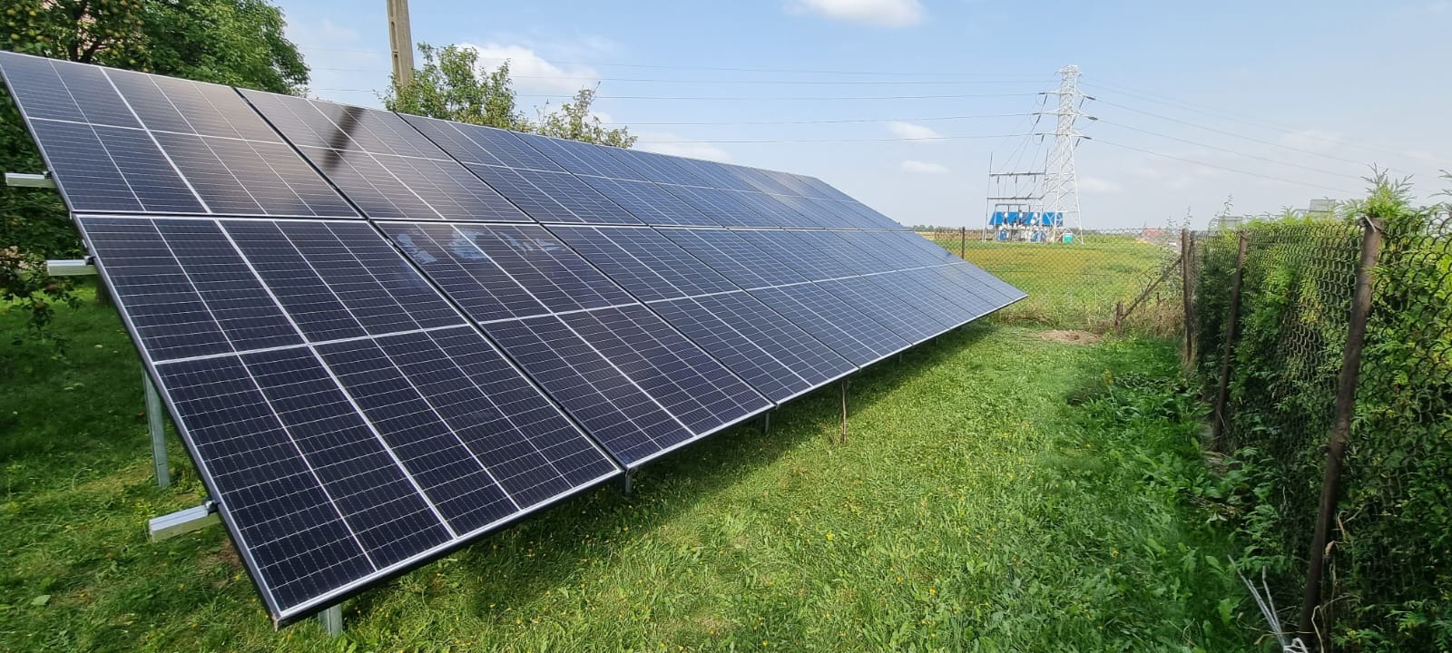 Rząd paneli słonecznych zamontowany na metalowej konstrukcji na trawiastym terenie, z widoczną linią wysokiego napięcia w oddali.