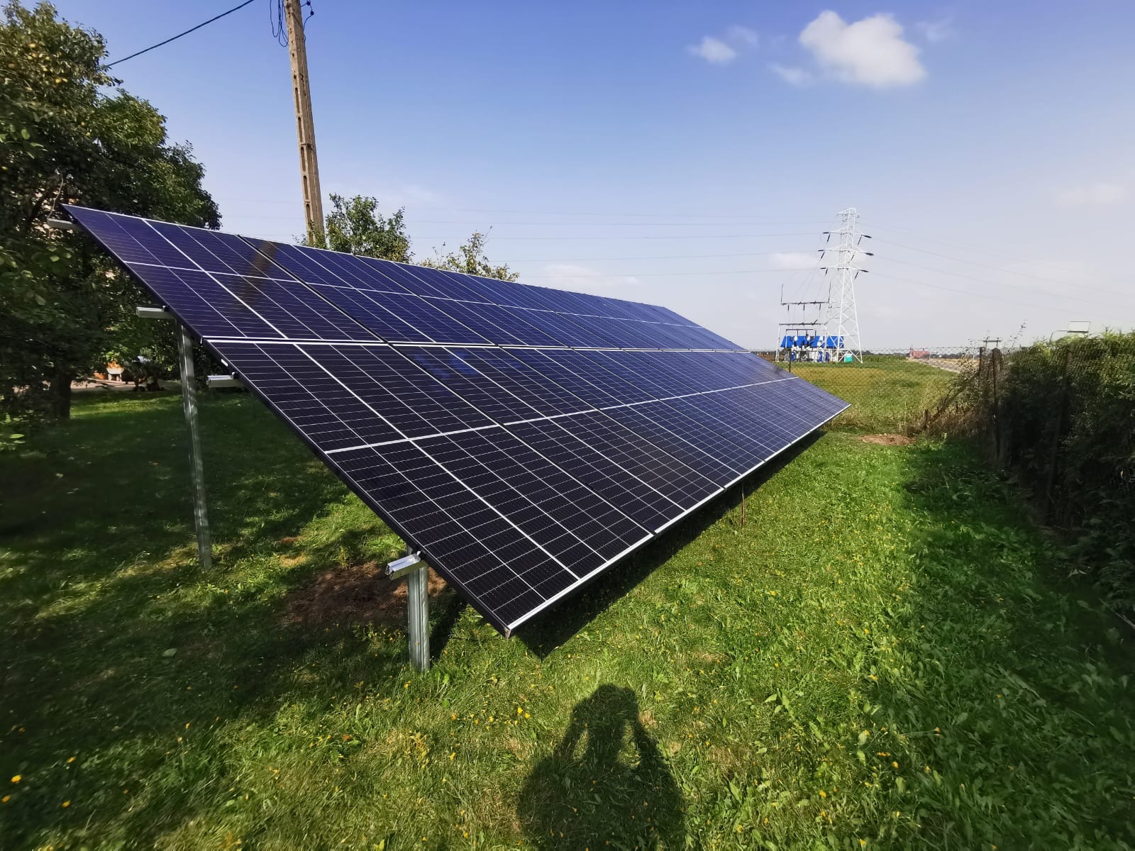Instalacja fotowoltaiczna na trawiastym terenie z widoczną linią wysokiego napięcia w tle, cień fotografa w dolnej części kadru.