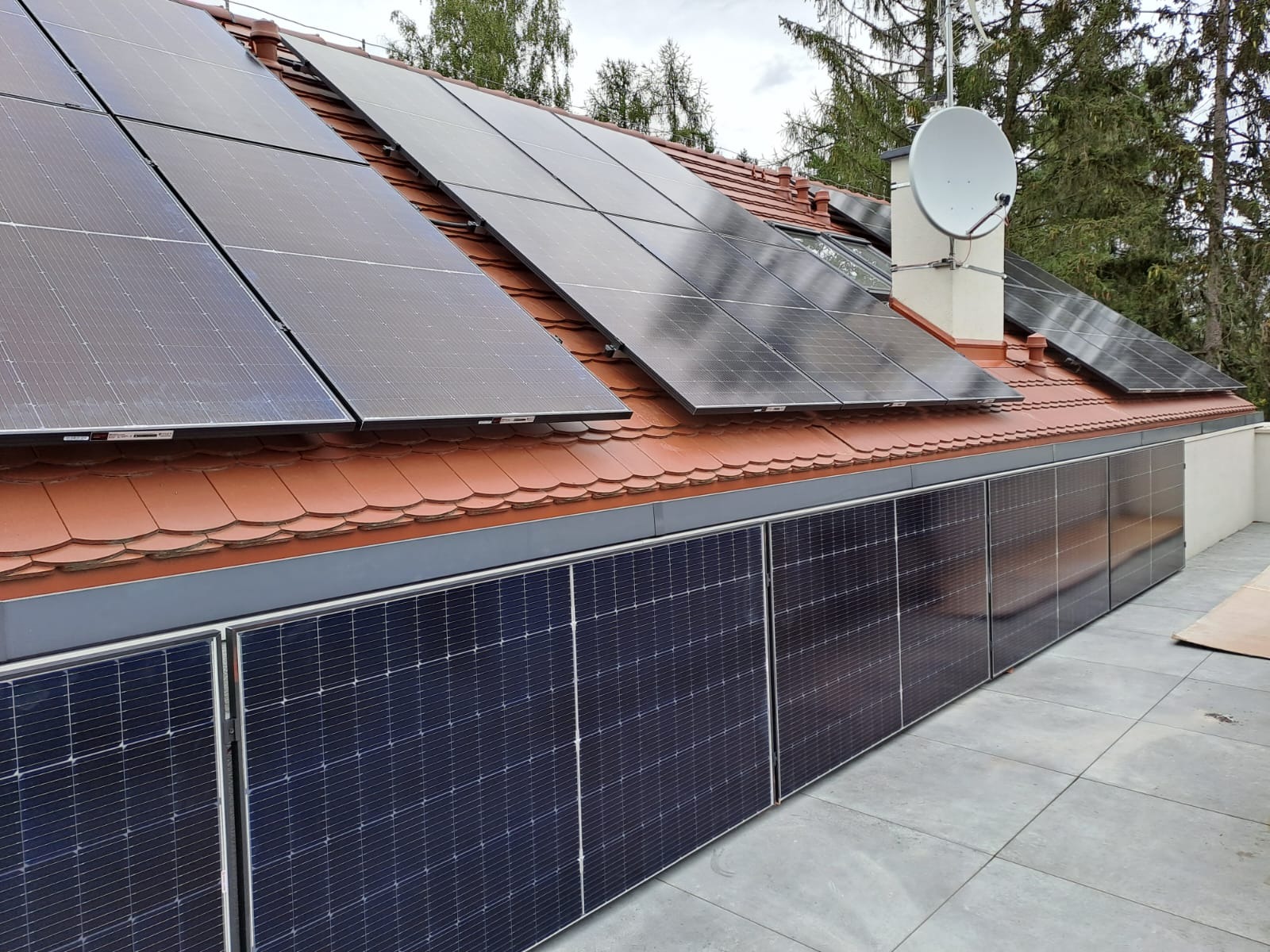 Panele słoneczne zamontowane na dachu z czerwonej dachówki i na ścianie budynku, obok komina z anteną satelitarną.