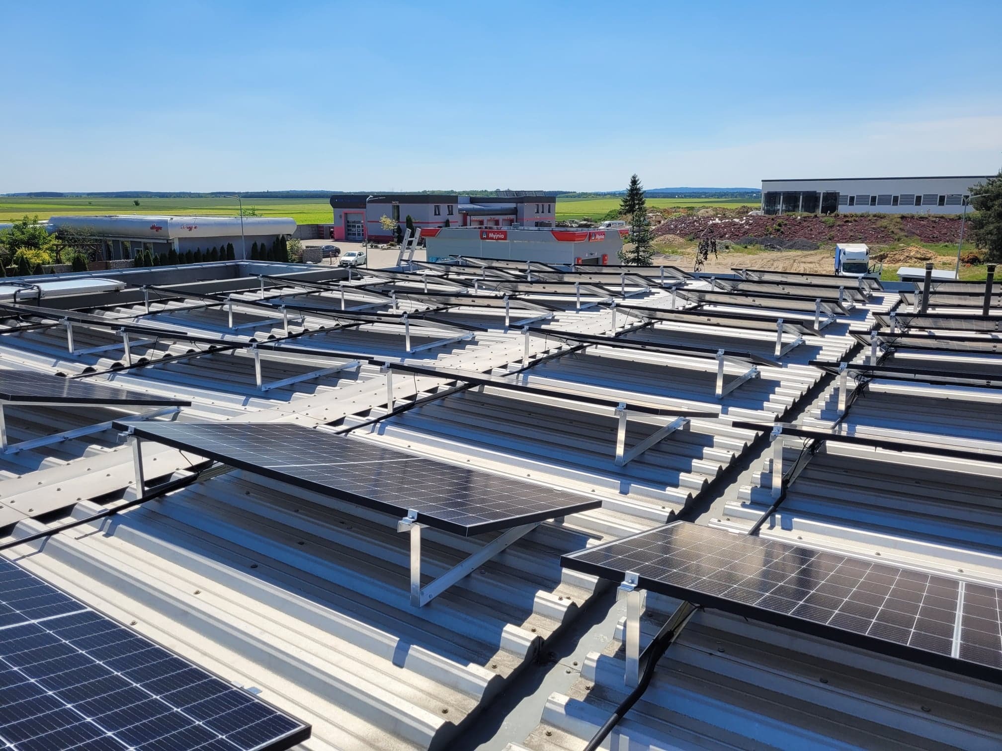 Montaż paneli fotowoltaicznych na dachu budynku komercyjnego, widoczne wsporniki i częściowo zainstalowane panele, w tle budynki Orlenu i zielone pola.