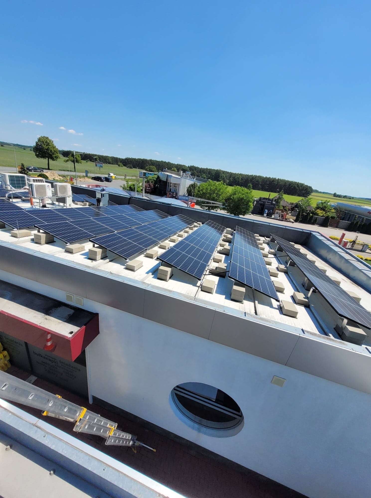 Instalacja paneli słonecznych na płaskim dachu budynku komercyjnego z widoczną drabiną i otoczeniem w słoneczny dzień.