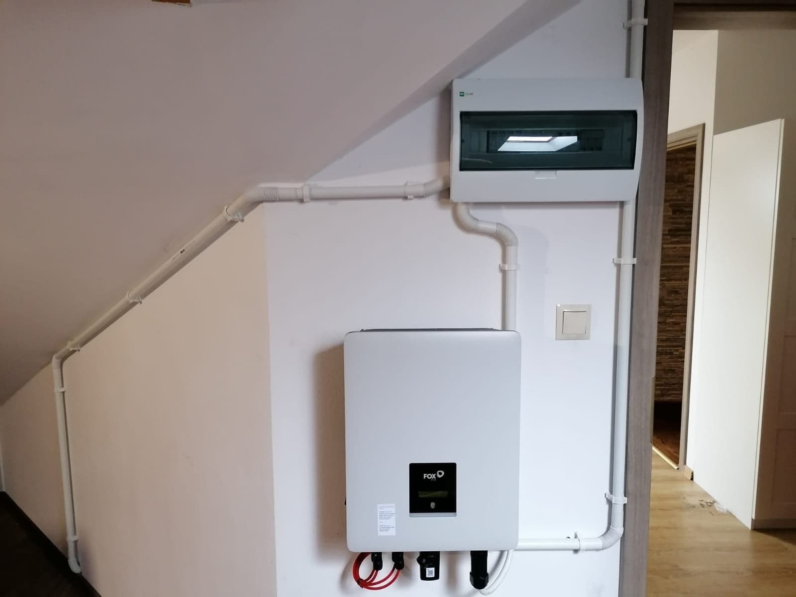 Biały inwerter fotowoltaiczny marki FOX zamontowany na ścianie pod skosem, z widoczną skrzynką rozdzielczą i okablowaniem w białych rurkach PCV.