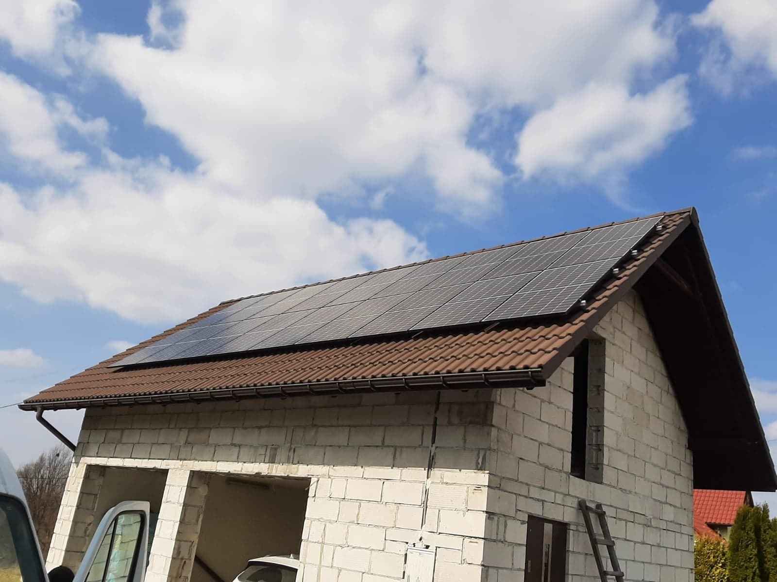 Panele słoneczne zamontowane na dachu domu w trakcie budowy w Bytomiu, widoczne białe bloczki i drabina oparta o ścianę, błękitne niebo z chmurami w tle.