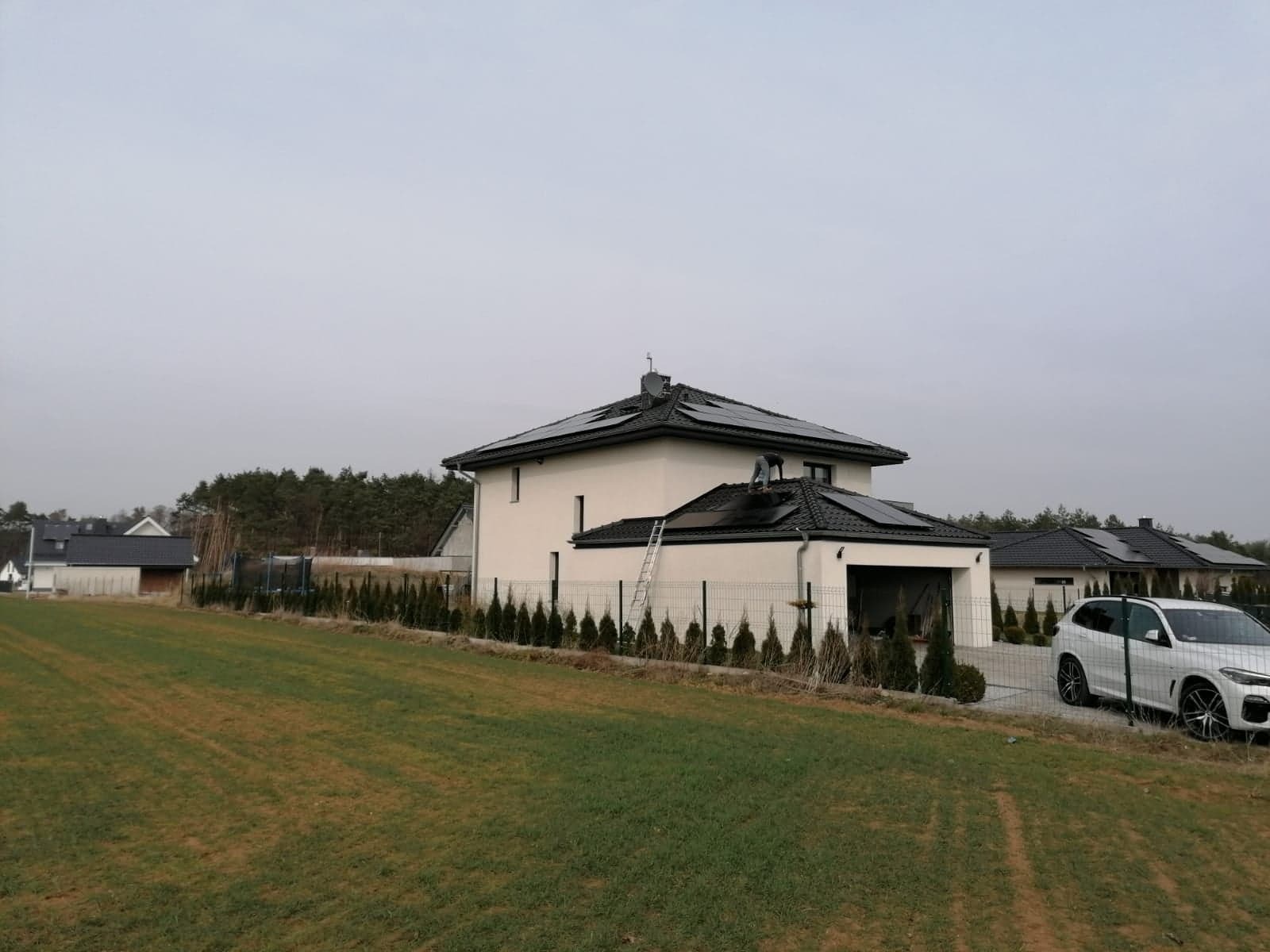 Montaż paneli słonecznych na dachu domu jednorodzinnego; widoczna drabina oparta o dach i instalator pracujący na panelach.