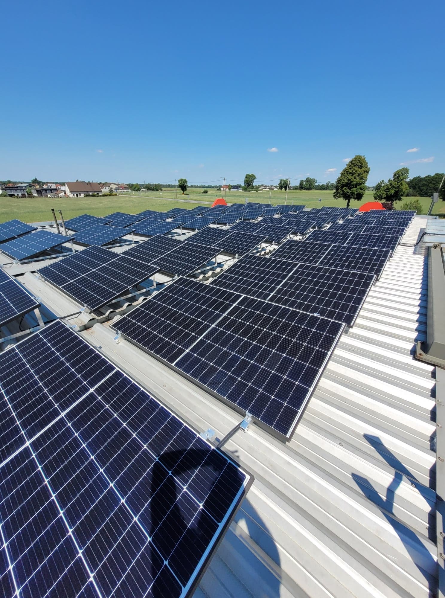 Rozległa instalacja paneli słonecznych na dachu budynku przemysłowego w słoneczny dzień, widoczne elementy mocujące i tło z zielonymi polami i budynkami.