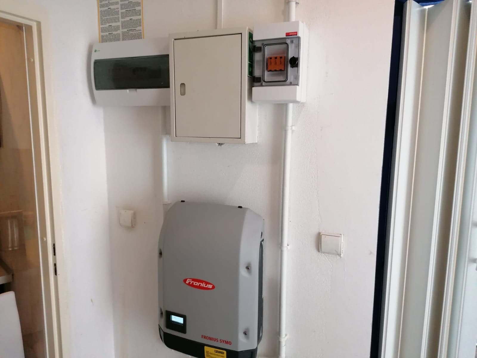 Szary inwerter Fronius Symo zamontowany na białej ścianie obok białych rur instalacyjnych i skrzynki elektrycznej z bezpiecznikami.