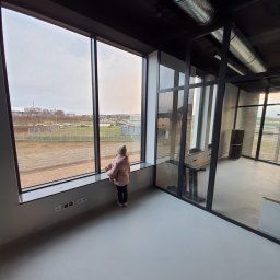 Hausenova SP.Zo.o - Wnętrze biura z dużym oknem, przez które widać krajobraz. Dziewczynka w różowej kurtce stoi przy oknie. Widoczne elementy wykończenia, szklane ścianki działowe i instalacja wentylacyjna pod sufitem.