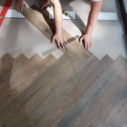 Hausenova SP.Zo.o - Montaż paneli podłogowych w jodełkę: widoczne dłonie układające panel, poziomica, nóż monterski i fragment ułożonej podłogi z ciemnego drewna.