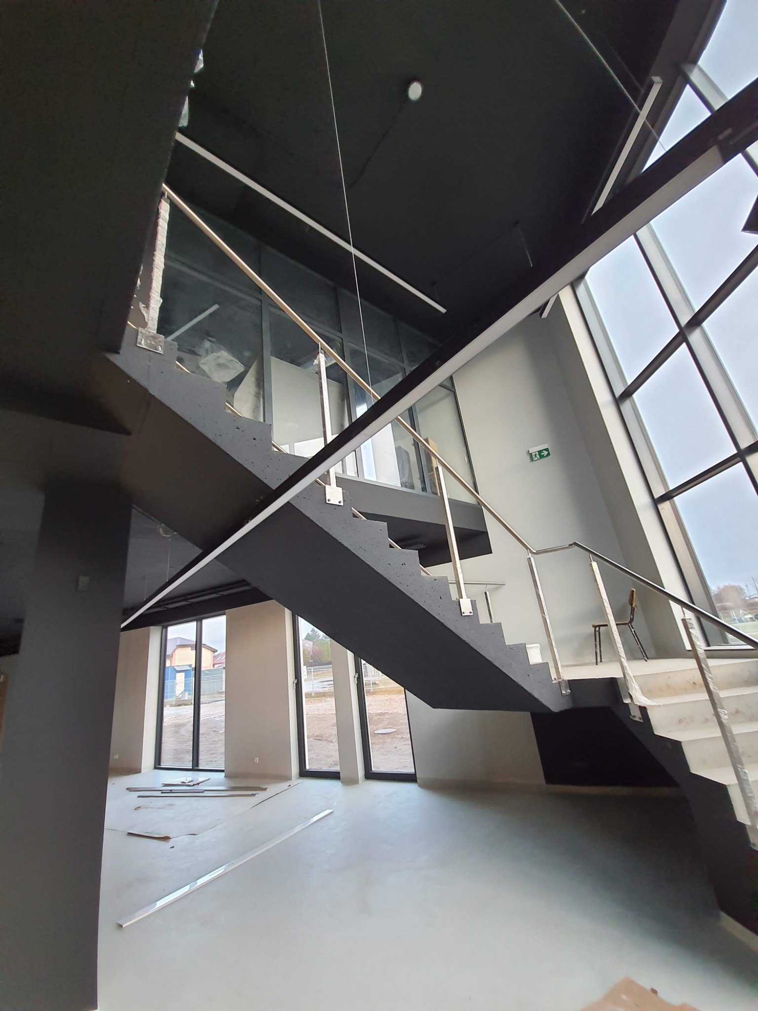 Wnętrze biura w trakcie wykańczania z widocznymi schodami z metalową balustradą, dużymi oknami i surowym betonem na podłodze.