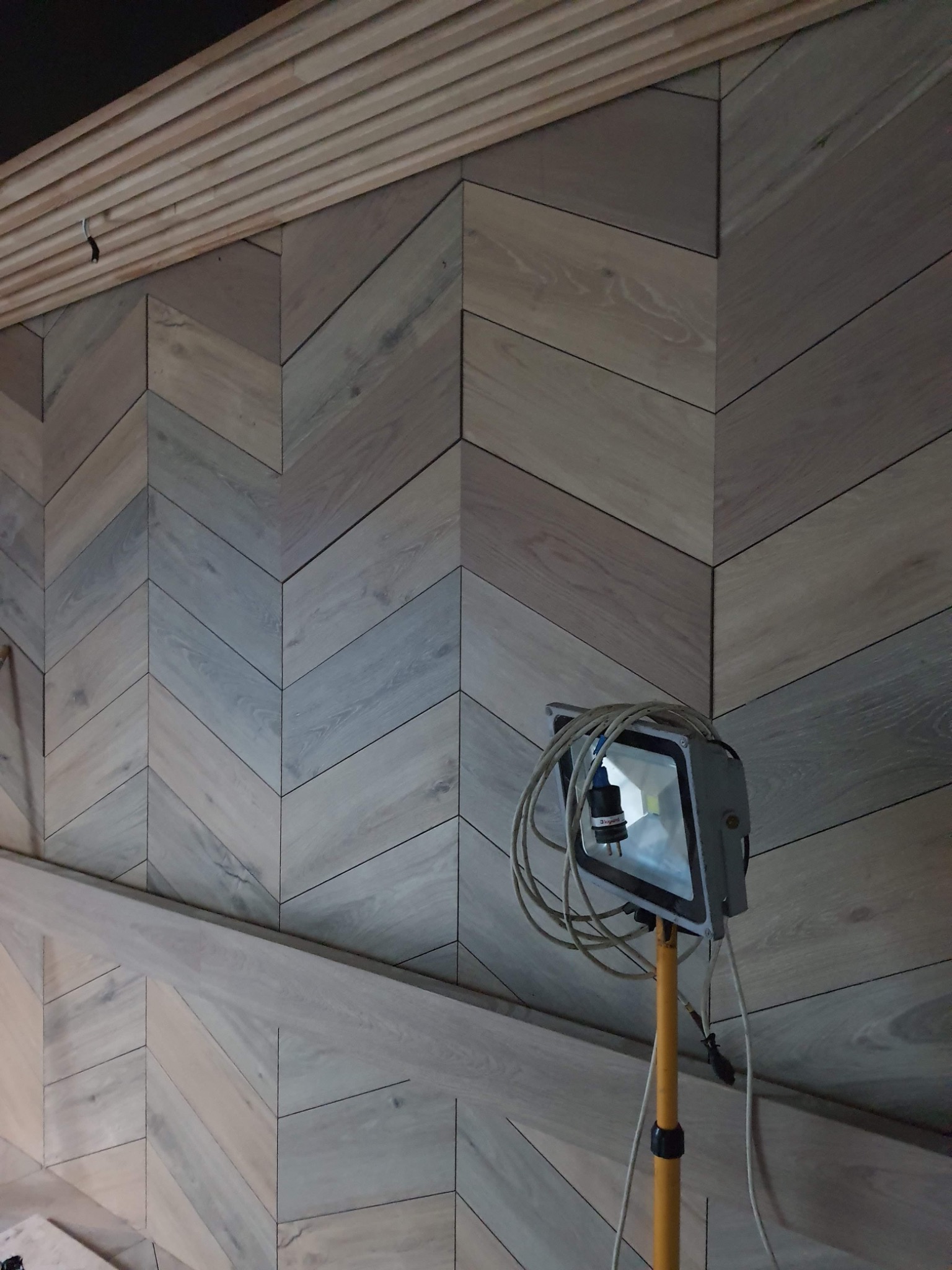 Ściana wykończona panelami drewnianymi ułożonymi we wzór jodełki, oświetlona lampą halogenową na żółtym statywie; widoczne również drewniane listwy na suficie.