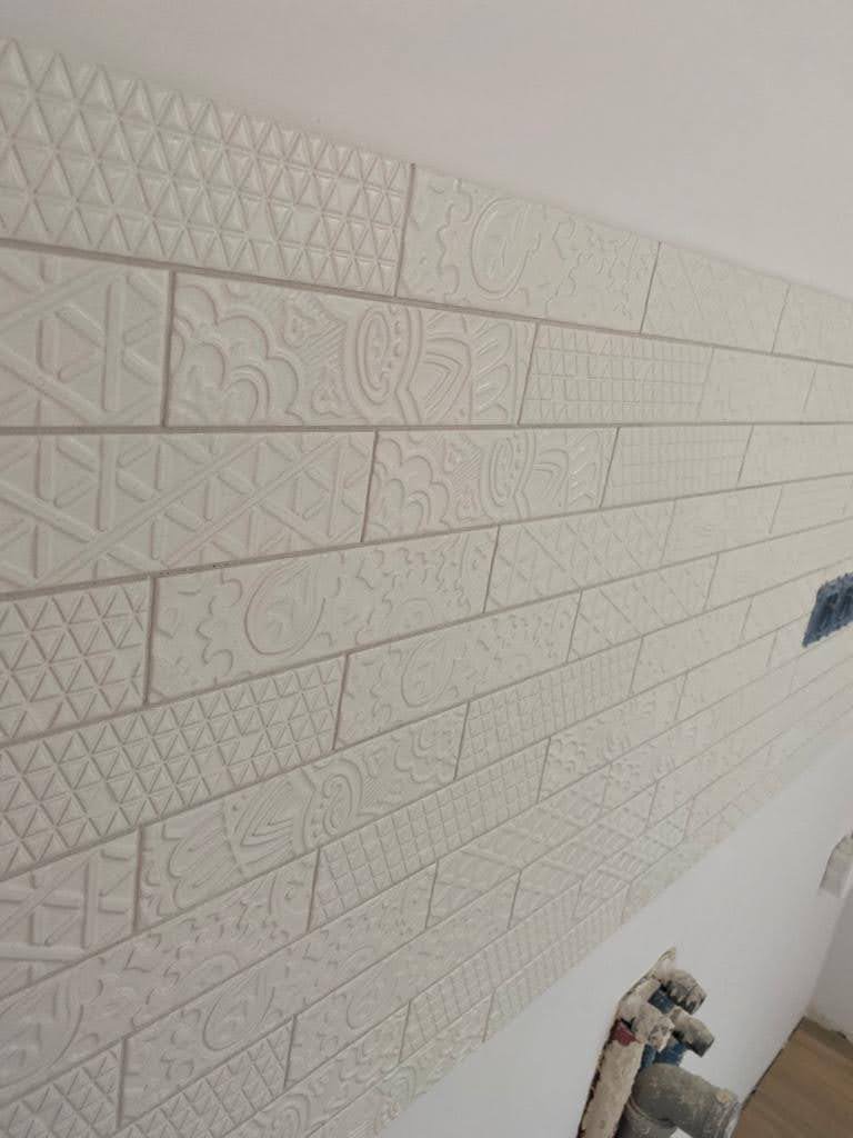 Ściana wyłożona białymi, podłużnymi płytkami z wytłaczanymi wzorami geometrycznymi i roślinnymi, ułożonymi horyzontalnie, widoczne fugi i fragment narzędzia glazurniczego w dolnym rogu.