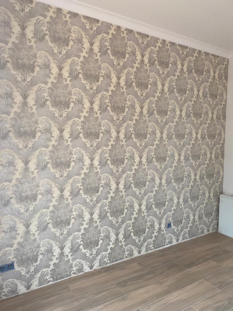 Ściana z szarą, wzorzystą tapetą w stylu retro, połączona z podłogą wyłożoną panelami imitującymi drewno, widoczny fragment białej listwy przypodłogowej i gniazdka elektryczne.
