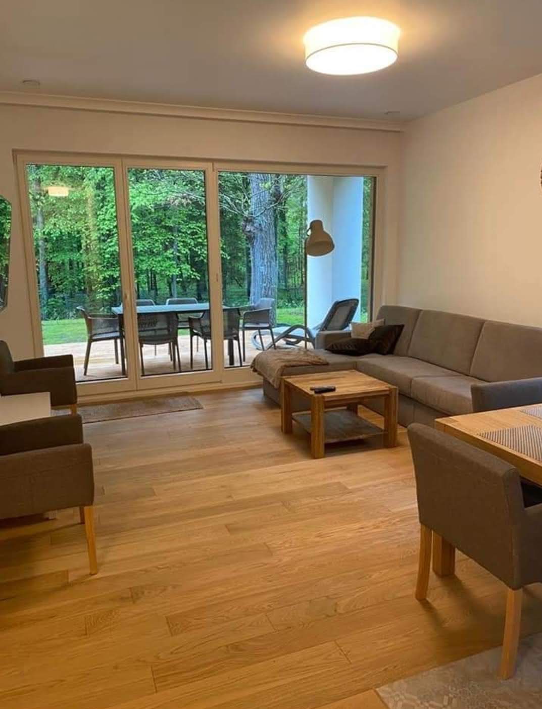 Jasny salon z drewnianą podłogą, dużymi przesuwnymi drzwiami balkonowymi wychodzącymi na zielony ogród i nowoczesnym oświetleniem sufitowym.