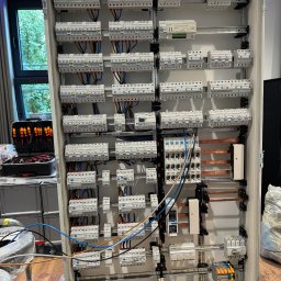 Kap-Bud - Rozdzielnica elektryczna w obudowie, widoczne liczne zabezpieczenia, przewody i szyny. Obok skrzynka z narzędziami i okno z widokiem na zieleń.