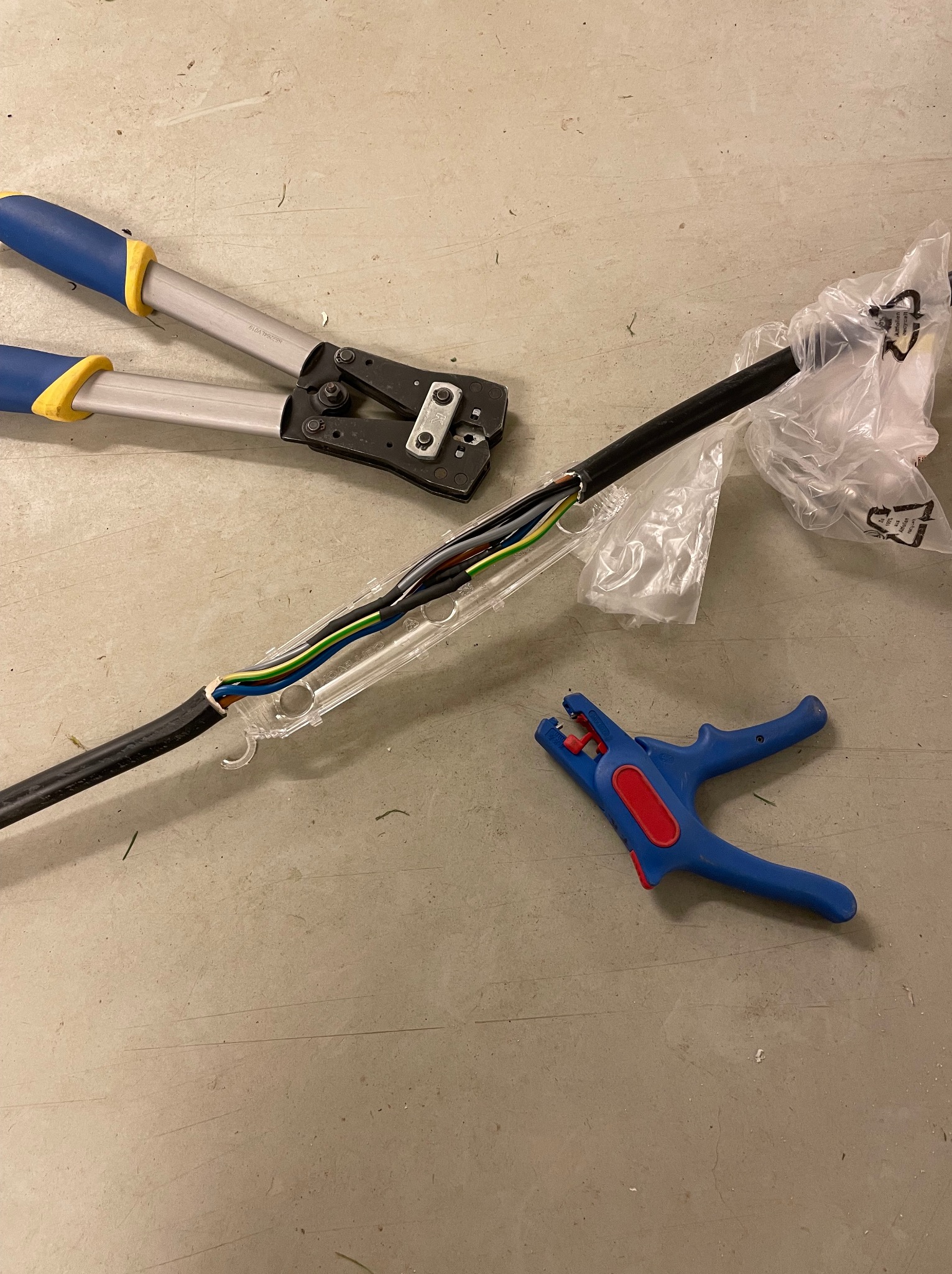 Obróbka kabla elektrycznego z użyciem narzędzi: zaciskarki z niebieskimi uchwytami i ściągacza izolacji. Kabel w przezroczystej osłonie, częściowo rozpakowany z folii.