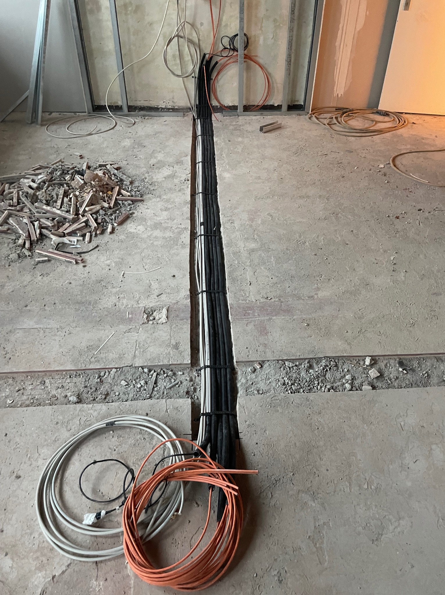 Wykop w posadzce z ułożonymi kablami elektrycznymi w czarnych peszlach, widoczne przewody pomarańczowe i białe, gruz i elementy konstrukcji szkieletowej.