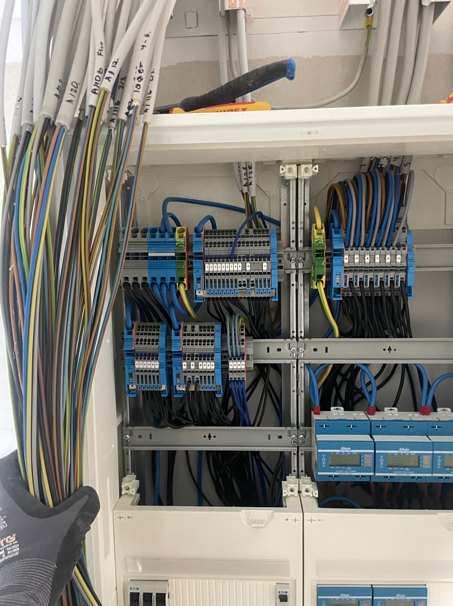 Rozdzielnica elektryczna z podłączonymi przewodami oznaczonymi etykietami, widoczne niebieskie moduły i szyny montażowe. Prace w toku, rękawica ochronna w kadrze.