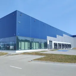 Nowoczesna hala magazynowa z niebiesko-szarą elewacją, przeszklonym biurem i rampami załadunkowymi w słoneczny dzień.