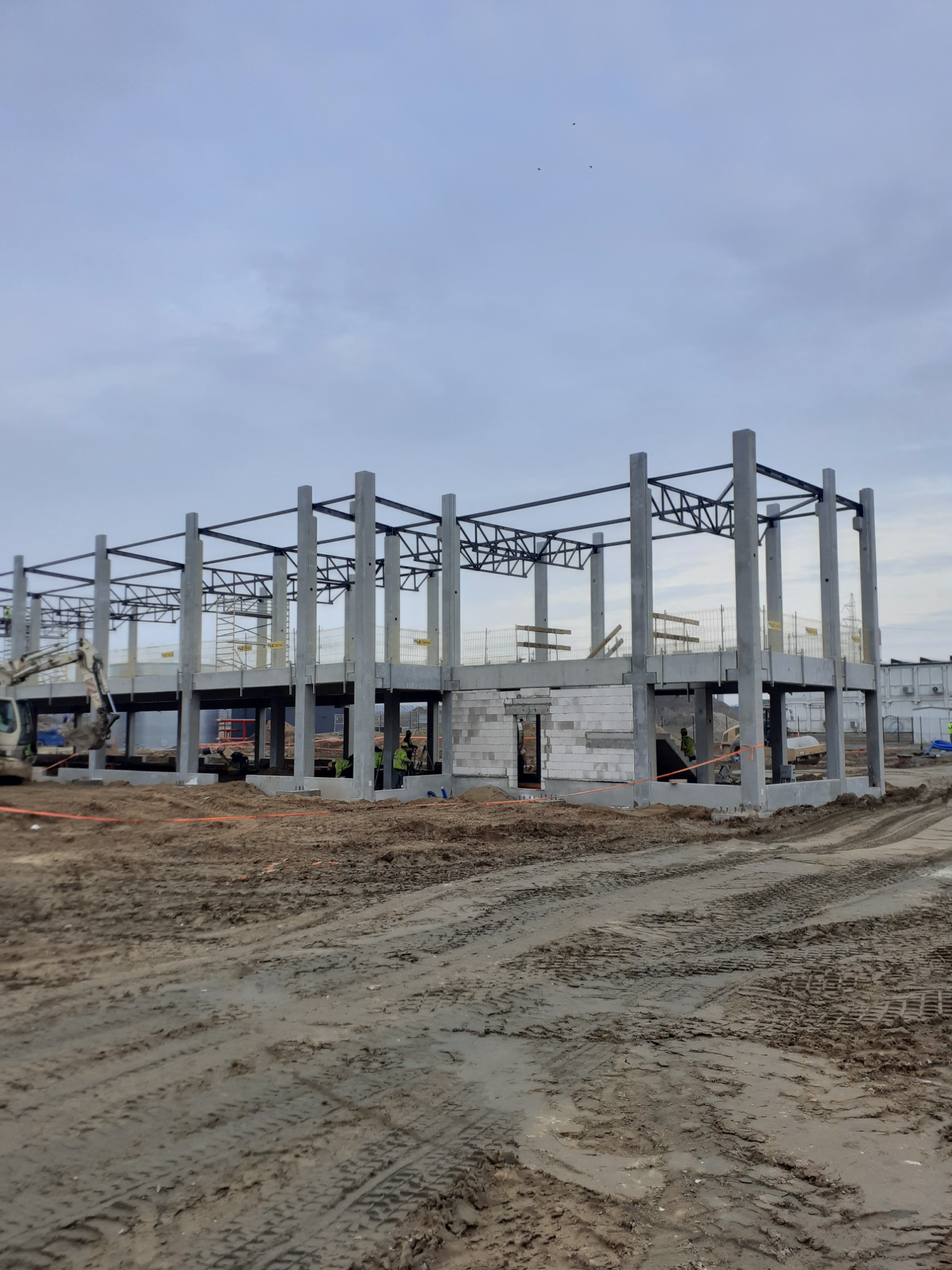Wznoszenie szkieletu hali z betonu i stali, widoczne fragmenty ścian z bloczków, ziemisty plac budowy z śladami opon, pochmurne niebo w tle.