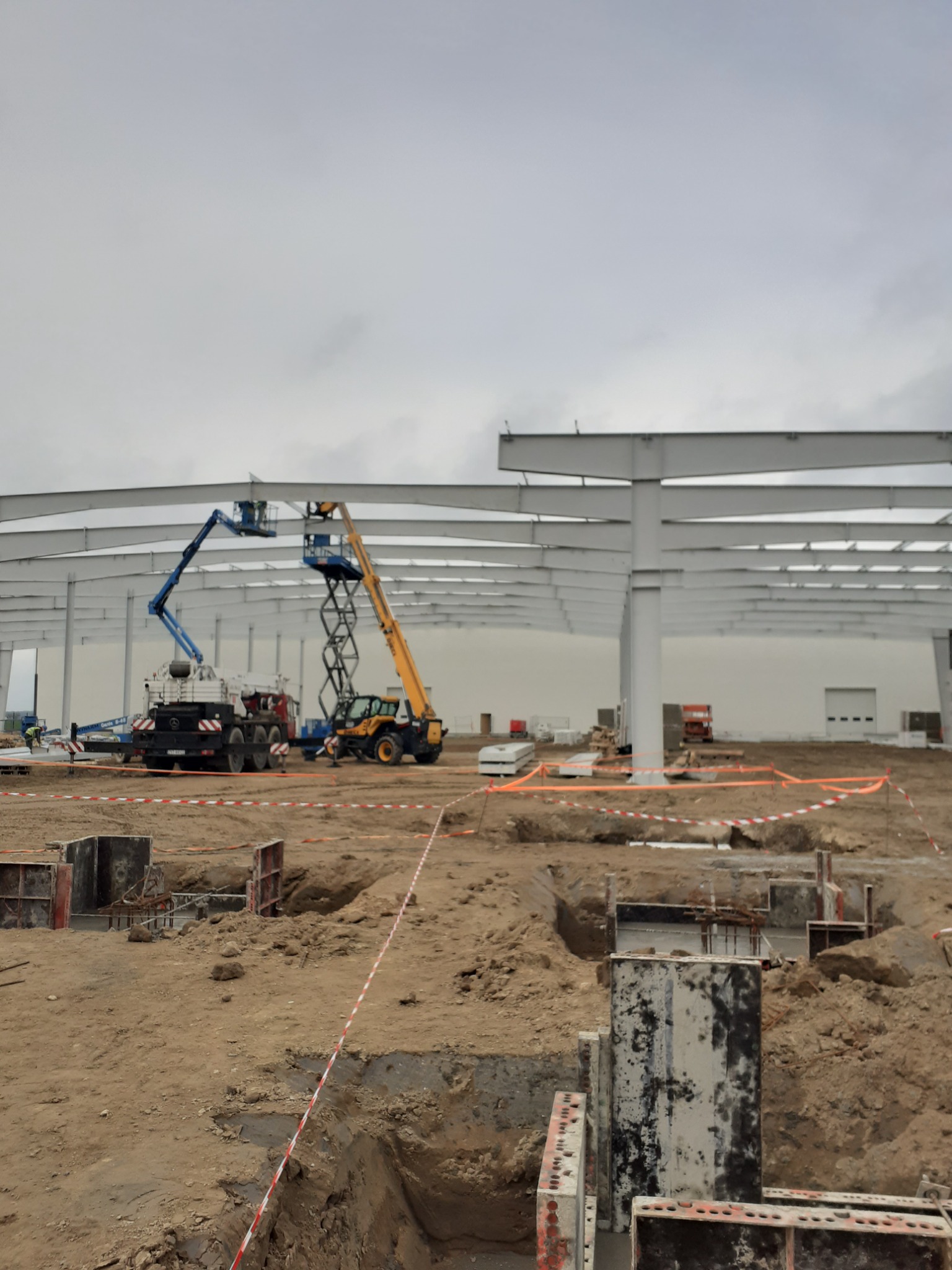 Wznoszenie konstrukcji stalowej hali: widoczne fundamenty, stalowe słupy, dźwigi platformowe i robotnicy na wysokościach.