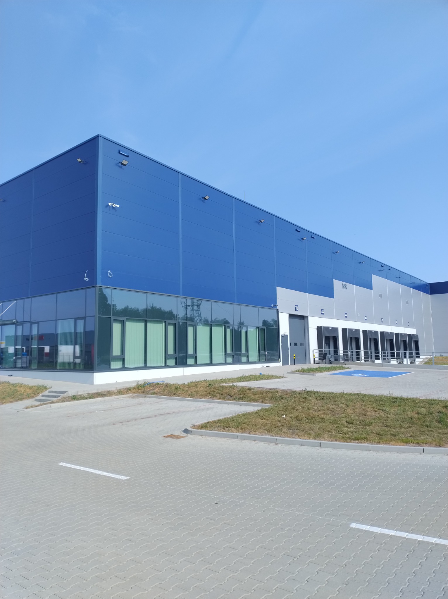 Nowoczesna hala magazynowa z niebiesko-szarą elewacją, przeszklonym biurem i rampami załadunkowymi w słoneczny dzień.