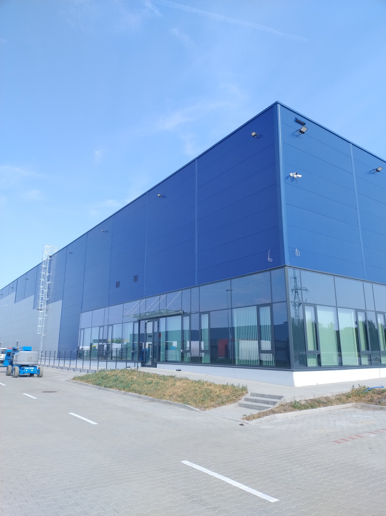 Nowoczesna hala magazynowa w kolorze niebieskim z przeszklonym biurem, widoczny podnośnik koszowy na zewnątrz.
