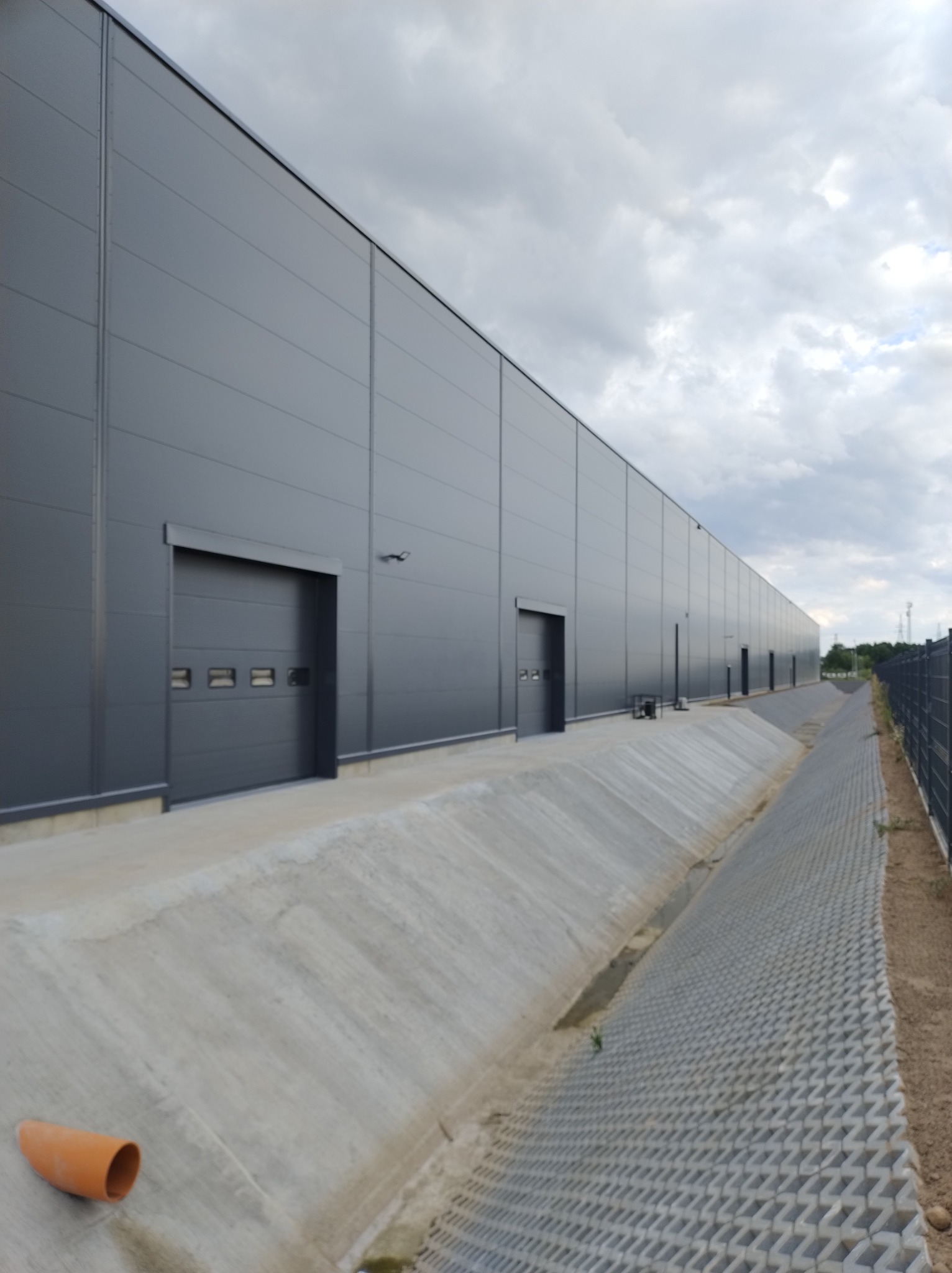Długa, szara hala przemysłowa z panelami warstwowymi i segmentowymi bramami garażowymi, widoczna z zewnątrz w pochmurny dzień. Obok hali betonowy rów odwadniający i ażurowa nawierzchnia.