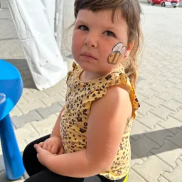 Dziewczynka z namalowaną na policzku pszczołą siedzi na żółtym krześle; w tle widać wóz strażacki.