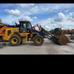 Żółto-czarna ładowarka teleskopowa JCB na placu budowy z innymi maszynami w tle, widok z boku, pochmurne niebo.