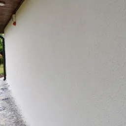 Świeżo otynkowana, jasna elewacja budynku jednorodzinnego z widoczną fakturą tynku baranka, zabezpieczone podłoże z resztkami zaprawy.