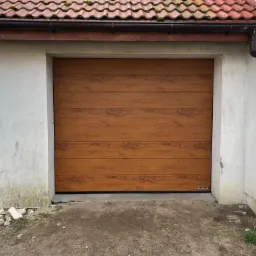 Zamknięta, segmentowa brama garażowa w kolorze drewna, z widocznym logo producenta w dolnym rogu, osadzona w surowym, betonowym otworze budynku z czerwoną dachówką.