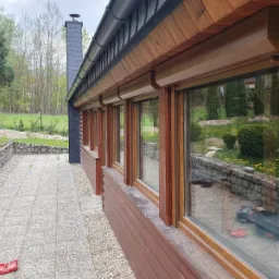 Dom z drewnianą elewacją i oknami z brązowymi roletami zewnętrznymi, widok z zewnątrz na kamienny chodnik i ogród.