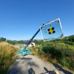 Mały żuraw pająk z przyssawkami podnosi duże okno na budowie, w tle dom i wzgórza, słoneczny dzień.