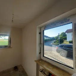 Wnętrze w trakcie remontu z dwoma oknami: mniejsze okno z widokiem na pole i większe okno z widokiem na ulicę z zaparkowanym samochodem. Na ramach okien widoczne naklejki producenta.