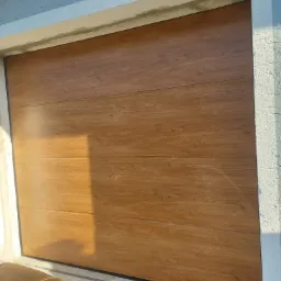 Zamknięta, segmentowa brama garażowa z paneli imitujących drewno, zamontowana w Nowej Rudzie, z widocznym logo producenta w prawym dolnym rogu.