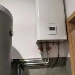 Biała pompa ciepła wisząca na ścianie z widocznymi rurami i zaworami oraz zbiornikiem na wodę obok, zamontowana w pomieszczeniu z szarą płytką na ścianie.