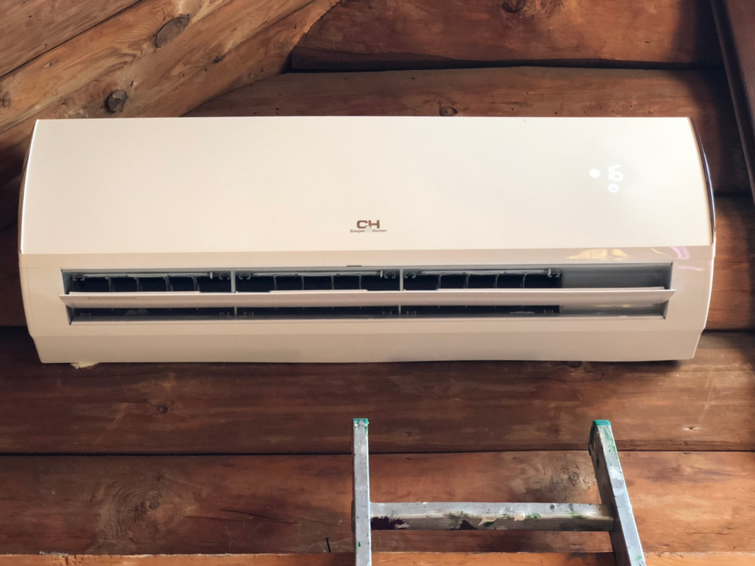 Biała jednostka klimatyzacji wewnętrznej marki Cooper&Hunter zamontowana na ścianie z drewnianych bali, widoczny fragment drabiny malarskiej.